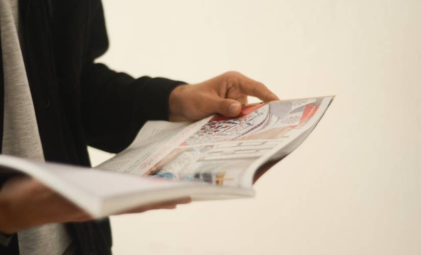 Kuvan vasemmassa reunassa seisova henkilö lukee lehteä. Henkilöstä näkyvät vain kädet, osa vatsaa ja osa vasenta käsivartta. Hän on pukeutunut mustaan ja harmaaseen ja pitelee käsissään avointa aikakauslehteä.